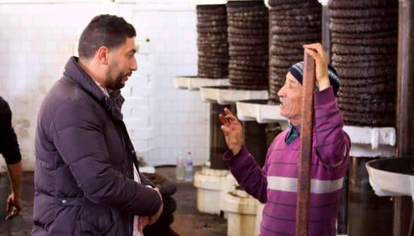 Two men talking inside an oil production facility, with equipment in the background. - Olive Oil Times