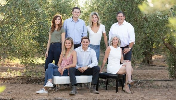 Seven individuals posing together outdoors in a natural setting with olive trees in the background. - Olive Oil Times