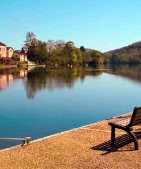 A wooden bench positioned along the edge of a calm river reflecting trees and buildings in the water. - Olive Oil Times