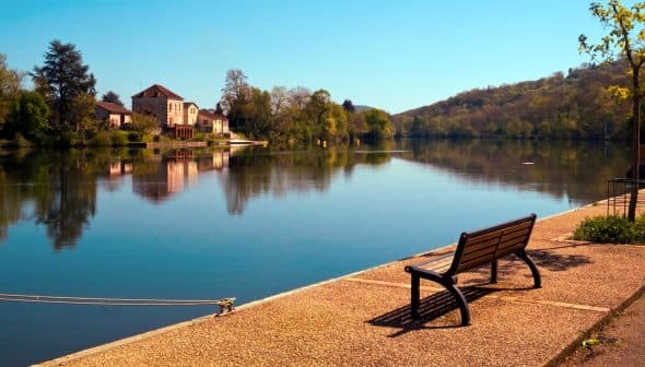 A wooden bench positioned along the edge of a calm river reflecting trees and buildings in the water. - Olive Oil Times