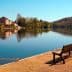 A wooden bench positioned along the edge of a calm river reflecting trees and buildings in the water. - Olive Oil Times