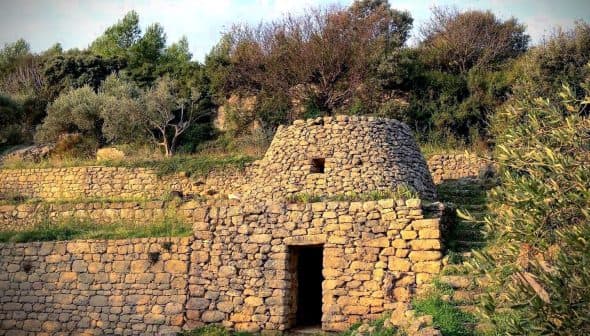 A stone structure with a conical roof surrounded by greenery and stone walls. - Olive Oil Times