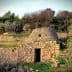 A stone structure with a conical roof surrounded by greenery and stone walls. - Olive Oil Times