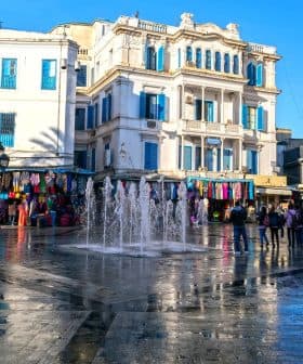 A fountain in a public square surrounded by buildings and people walking. - Olive Oil Times