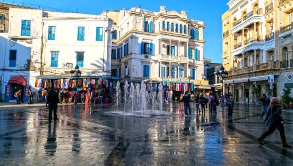A fountain in a public square surrounded by buildings and people walking. - Olive Oil Times