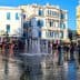 A fountain in a public square surrounded by buildings and people walking. - Olive Oil Times