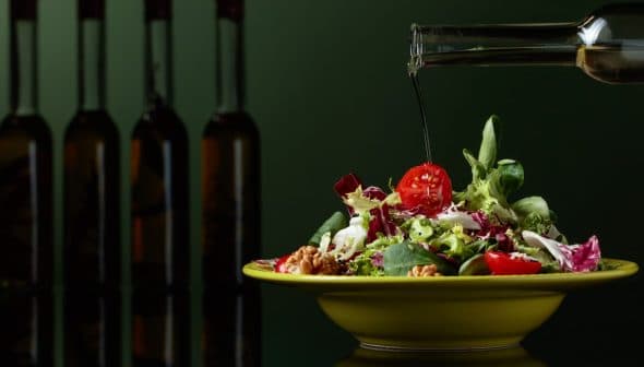 Olive oil being poured over a fresh salad in a yellow bowl with various vegetables. - Olive Oil Times