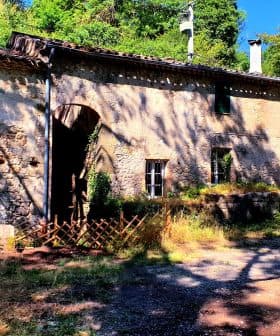 A rustic stone building featuring green windows and a door, surrounded by greenery. - Olive Oil Times