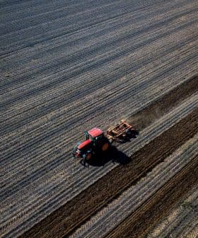 Red tractor tilling soil in a large agricultural field with distinct rows. - Olive Oil Times