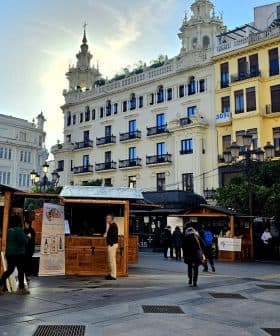 Wooden market stalls set up in an urban square with buildings in the background. - Olive Oil Times