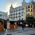 Wooden market stalls set up in an urban square with buildings in the background. - Olive Oil Times