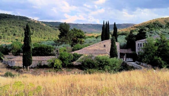 A stone building with a tiled roof, surrounded by trees and tall grass in a rural landscape. - Olive Oil Times
