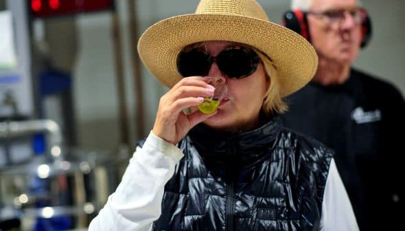 Woman wearing a straw hat and sunglasses tasting olive oil in a factory setting. - Olive Oil Times