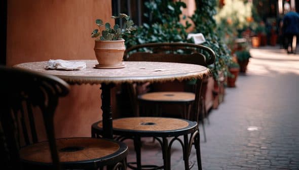 A round table with a potted plant and three chairs in a narrow outdoor setting. - Olive Oil Times