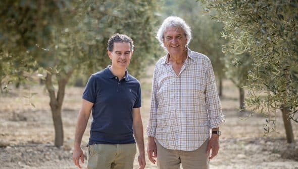 Two men standing in an olive grove, both smiling and dressed casually in light clothing. - Olive Oil Times