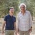 Two men standing in an olive grove, both smiling and dressed casually in light clothing. - Olive Oil Times