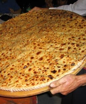 A person holding a large round flatbread topped with seeds and spices, presented on a wooden tray. - Olive Oil Times