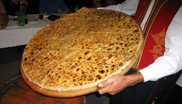 A person holding a large round flatbread topped with seeds and spices, presented on a wooden tray. - Olive Oil Times