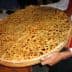 A person holding a large round flatbread topped with seeds and spices, presented on a wooden tray. - Olive Oil Times