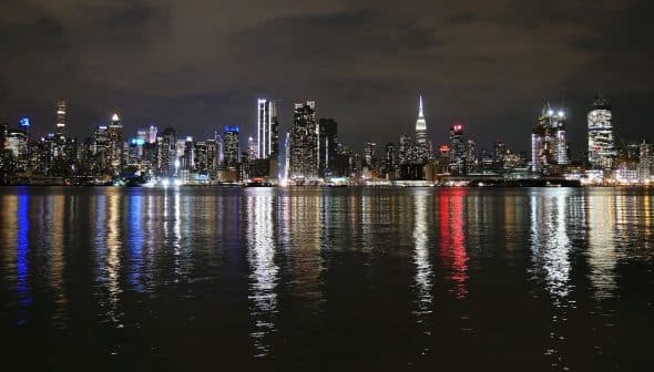 Night view of the New York City skyline with illuminated buildings reflecting on the water. - Olive Oil Times