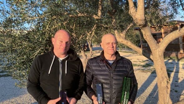 Two men standing outdoors holding bottles of olive oil in front of an olive tree. - Olive Oil Times