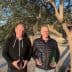 Two men standing outdoors holding bottles of olive oil in front of an olive tree. - Olive Oil Times