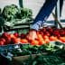 A hand reaching for ripe red tomatoes at a market stall filled with fresh vegetables. - Olive Oil Times