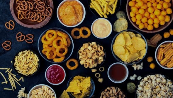 An assortment of snack foods including chips, pretzels, and popcorn arranged in bowls on a dark surface. - Olive Oil Times