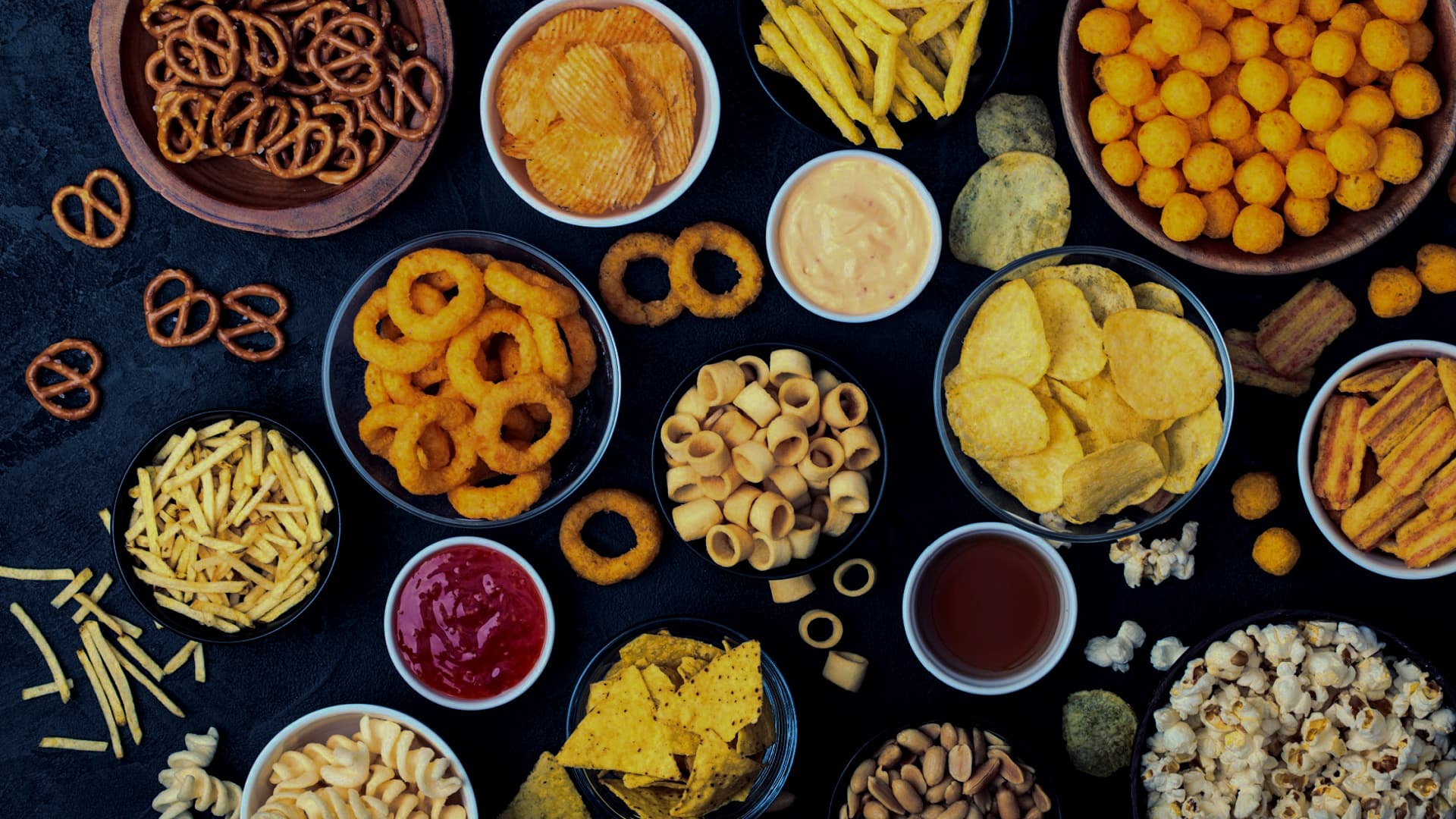 An assortment of snack foods including chips, pretzels, and popcorn arranged in bowls on a dark surface. - Olive Oil Times