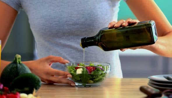 Individual pouring olive oil from a dark glass bottle onto a fresh salad in a clear bowl. - Olive Oil Times