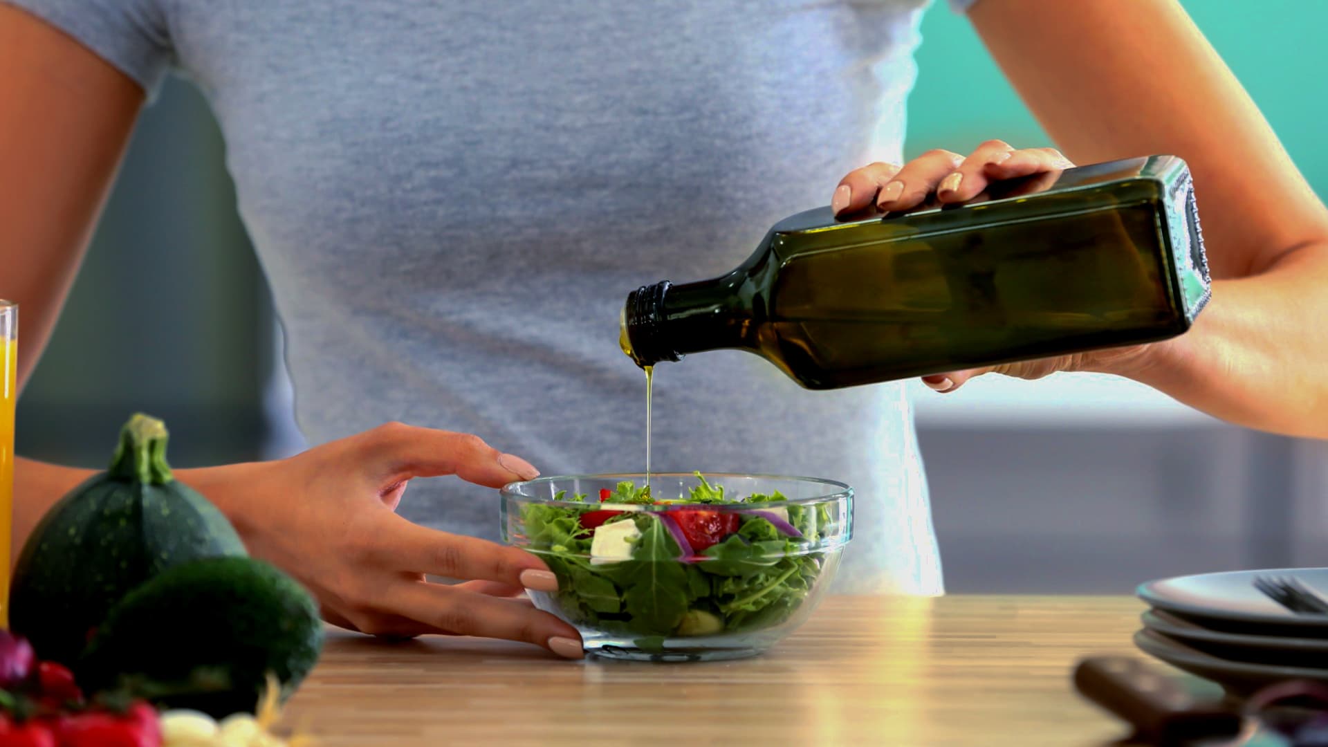 Individual pouring olive oil from a dark glass bottle onto a fresh salad in a clear bowl. - Olive Oil Times