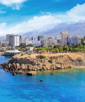 Coastal view featuring a city skyline with mountains in the background and clear blue water. - Olive Oil Times