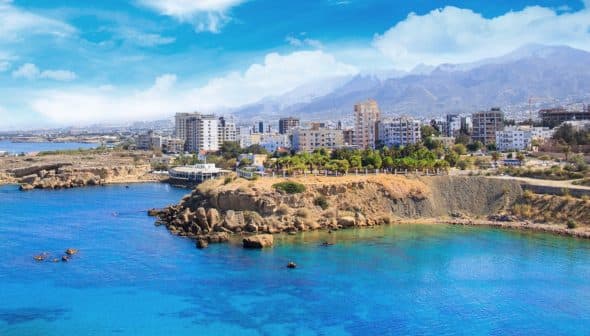 Coastal view featuring a city skyline with mountains in the background and clear blue water. - Olive Oil Times