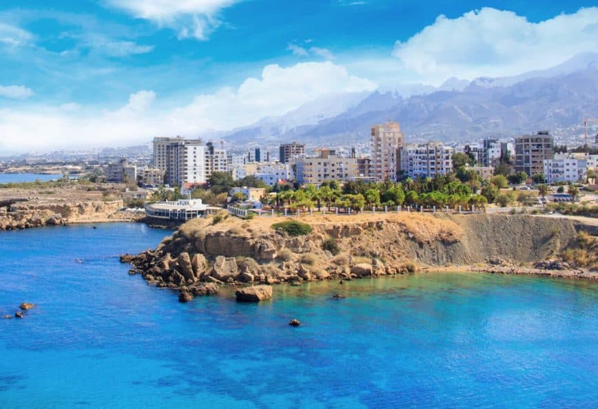 Coastal view featuring a city skyline with mountains in the background and clear blue water. - Olive Oil Times