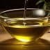A clear glass bowl filled with olive oil, with olive branches placed nearby on a wooden surface. - Olive Oil Times