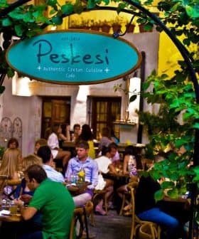 Entrance sign of Peskesi restaurant with greenery and patrons dining inside. - Olive Oil Times