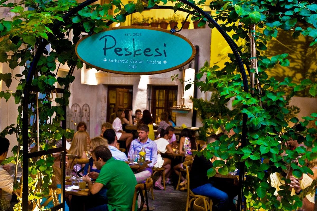 Entrance sign of Peskesi restaurant with greenery and patrons dining inside. - Olive Oil Times