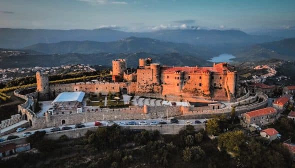 Aerial view of a historic castle surrounded by mountains and greenery. - Olive Oil Times