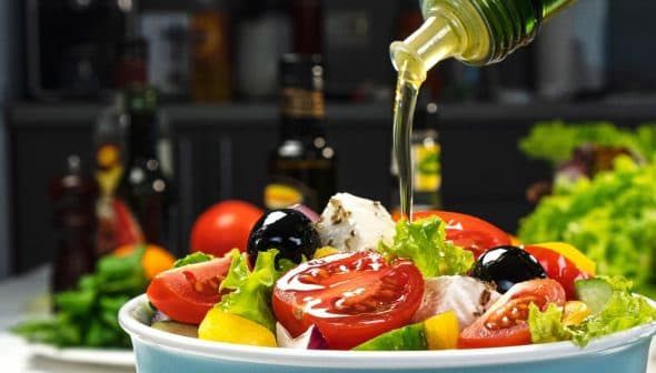 A bowl of fresh salad being drizzled with olive oil, featuring tomatoes, lettuce, and olives. - Olive Oil Times
