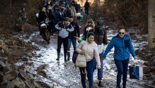A group of people walking along a muddy path in a wooded area, some carrying bags. - Olive Oil Times