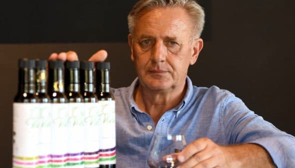 Man with gray hair holding a glass in front of a row of olive oil bottles on a table. - Olive Oil Times