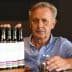 Man with gray hair holding a glass in front of a row of olive oil bottles on a table. - Olive Oil Times