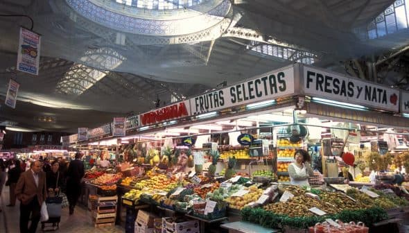 Market stall displaying a variety of fruits and vegetables under a large ceiling structure. - Olive Oil Times