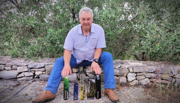 A man seated on a stone wall with various olive oil bottles displayed in front of him. - Olive Oil Times