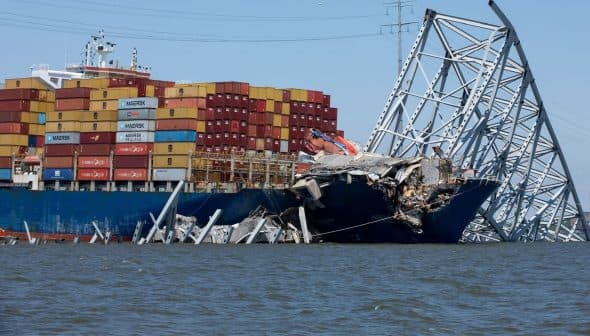 Cargo ship with shipping containers partially submerged and damaged, leaning against a collapsed structure. - Olive Oil Times