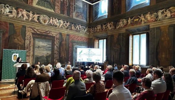 Audience seated in a historic hall during a conference presentation with decorative walls and a large screen. - Olive Oil Times