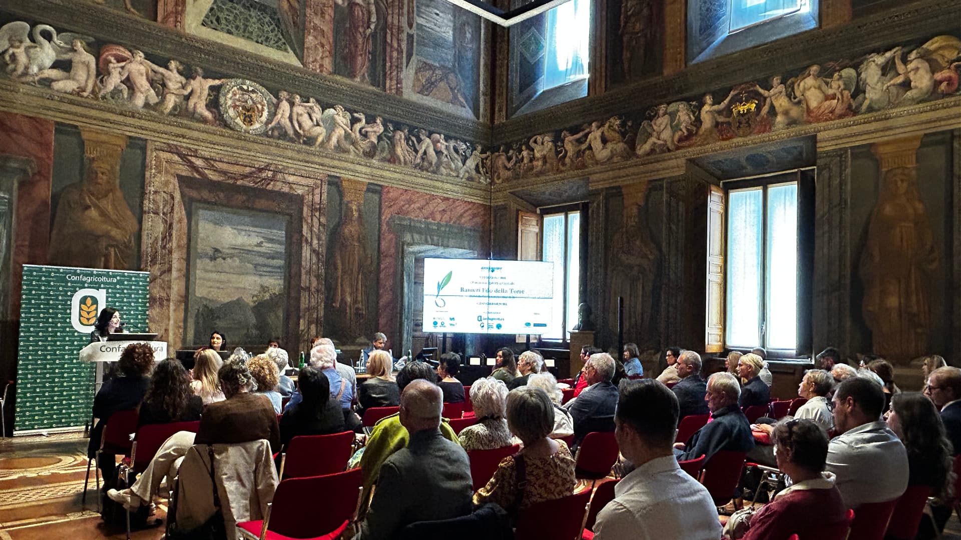 Audience seated in a historic hall during a conference presentation with decorative walls and a large screen. - Olive Oil Times