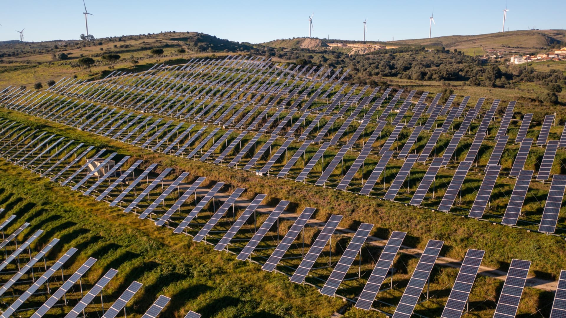 Aerial view of solar panels arranged in rows on a hillside with wind turbines in the background. - Olive Oil Times