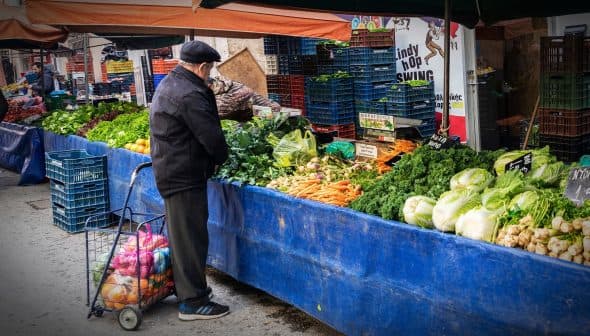 Man examining fresh vegetables at a market stall with various produce displayed. - Olive Oil Times
