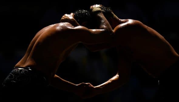 Two wrestlers in a close hold during a match, with their bodies glistening under the light. - Olive Oil Times